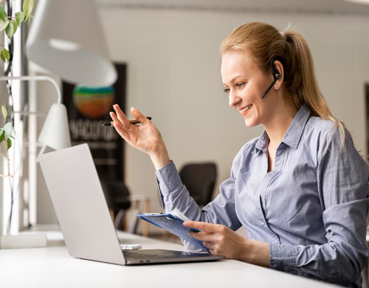 Mulher sorridente trabalhando como assistente virtual, usando fones de ouvido com microfone enquanto conversa com um cliente e faz anotações em frente ao laptop.
