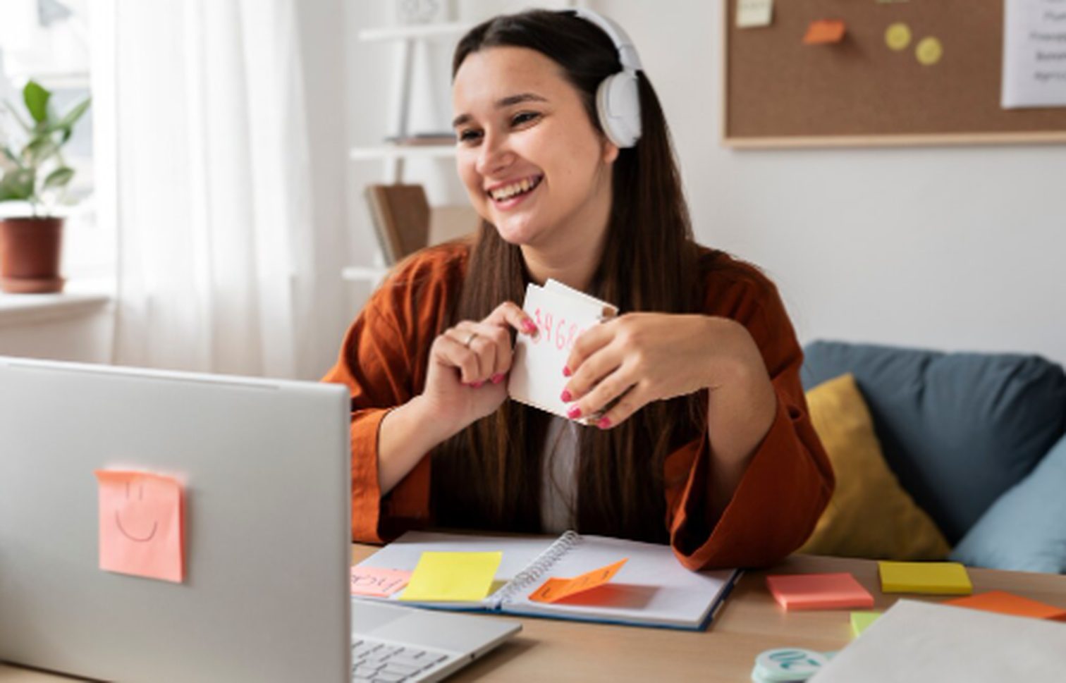 Professora sorridente ministrando aulas particulares online com fones de ouvido, mostrando cartões educativos diante do notebook em um ambiente de estudo organizado.