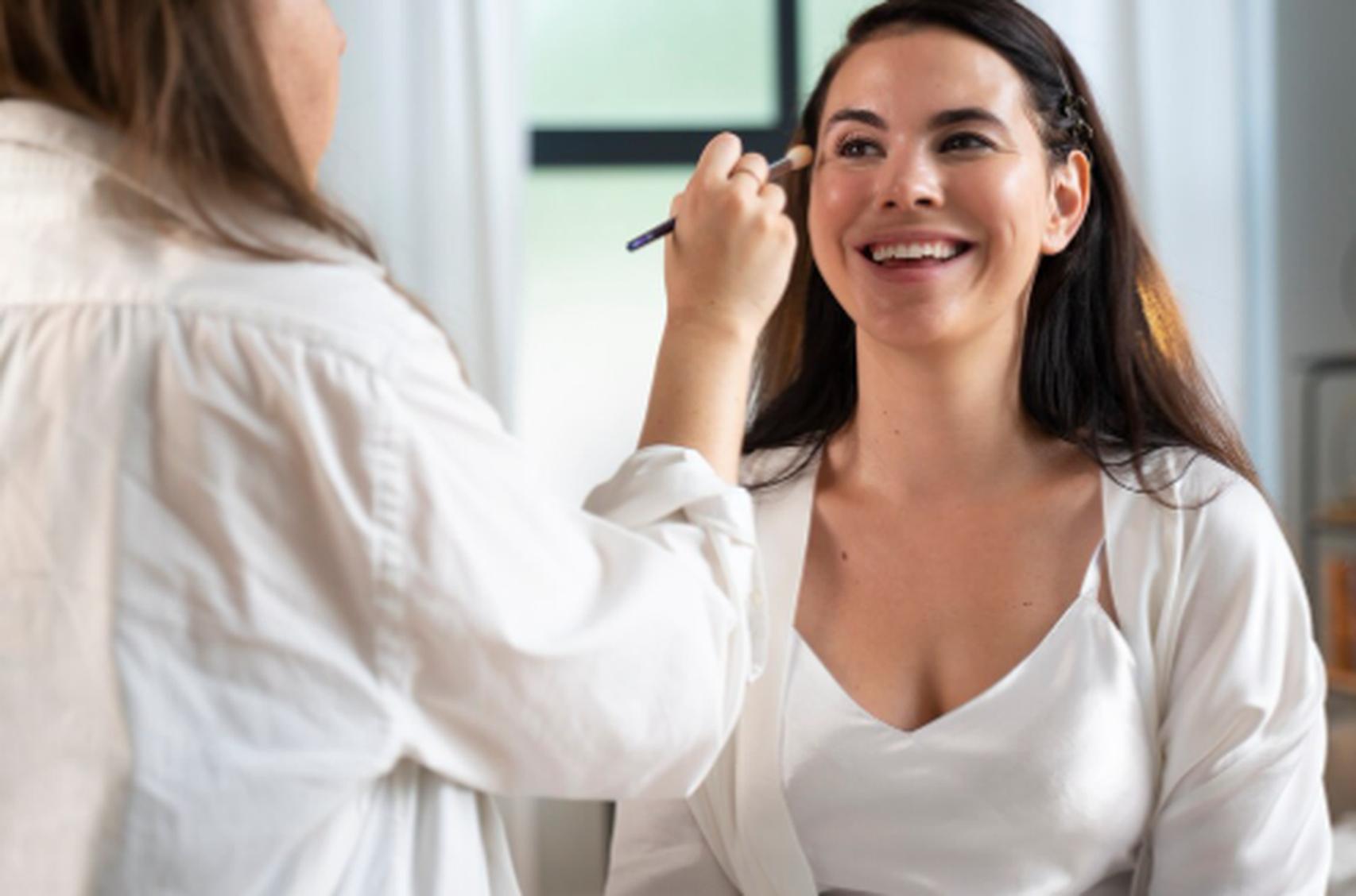Mulher sorridente recebe maquiagem profissional em ambiente iluminado, representando os serviços de estética e beleza em casa com praticidade, conforto e qualidade no atendimento personalizado.