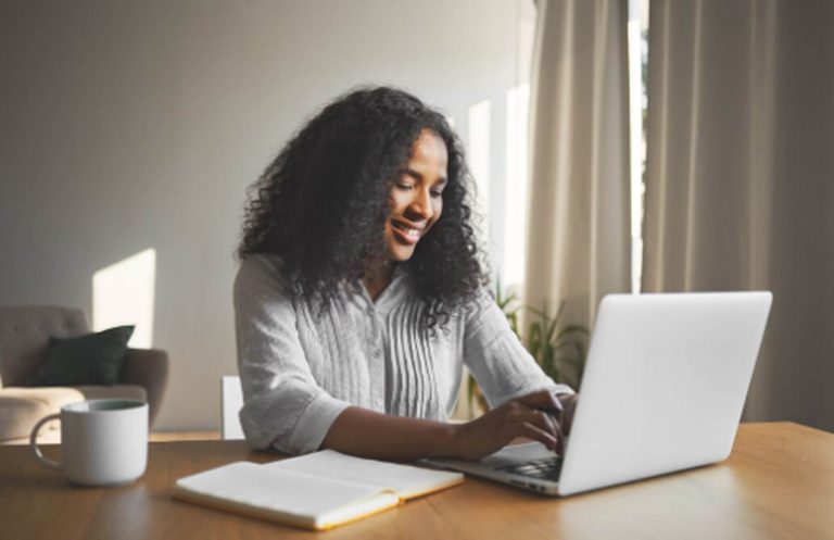 Uma mulher sorridente trabalha em um laptop em uma mesa iluminada pela luz natural, com um caderno e uma xícara ao lado, representando a rotina produtiva e inspiradora de uma redatora freelancer em seu espaço de trabalho.