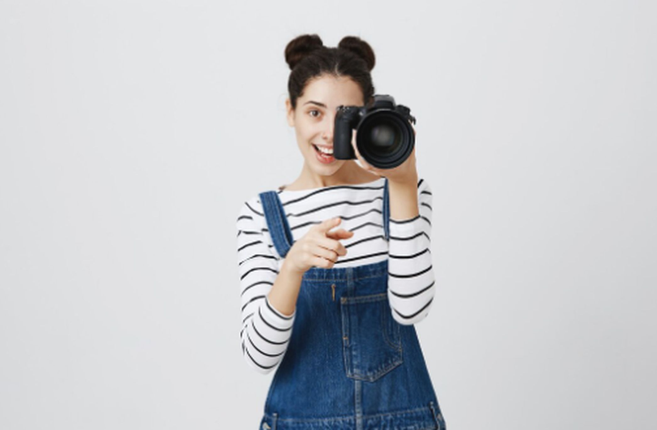 Mulher jovem sorridente, vestindo jardineira jeans e blusa listrada, segura uma câmera profissional apontada para frente em um estúdio claro, representando serviços de fotografia modernos e criativos.