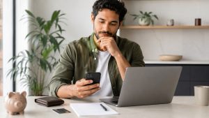 Homem sentado em uma mesa organizada, segurando um celular enquanto analisa informações financeiras em um notebook. Sobre a mesa há um cofrinho, uma carteira, um cartão bancário, uma folha em branco e uma caneta, sugerindo planejamento, comparação de opções e cuidado antes de solicitar um empréstimo pessoal. O ambiente é moderno, claro e minimalista, com plantas ao fundo e luz natural suave.
